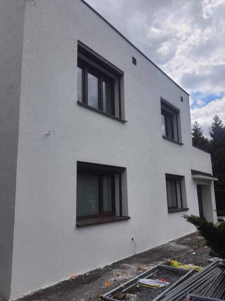 Dwukondygnacyjny budynek z nową, jasną elewacją, ciemnymi ramami okiennymi i zewnętrznymi roletami, widoczne rusztowanie na ziemi.