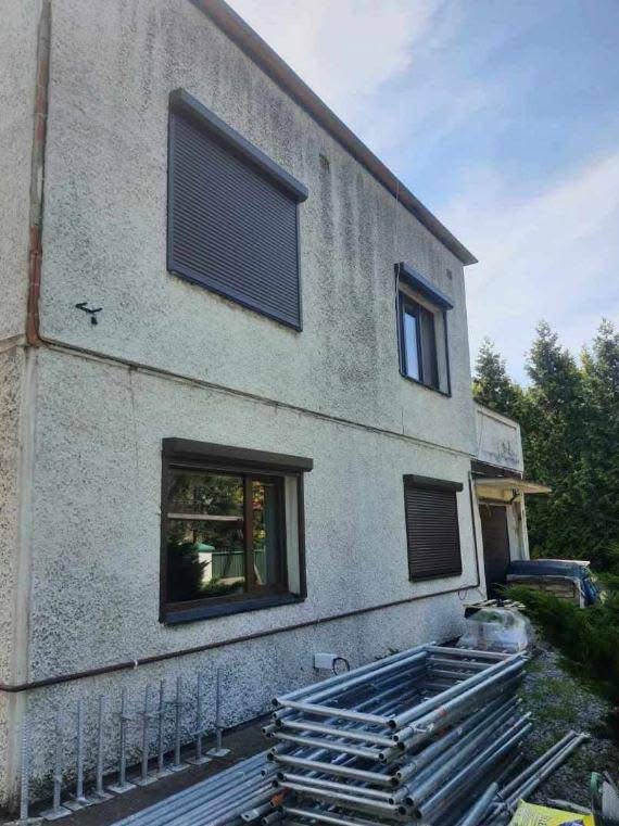 Dwupiętrowy dom z elewacją o szorstkiej fakturze, z trzema oknami z brązowymi roletami, stos rusztowań leżący u podstawy ściany, widok z dołu na elewację w pochmurny dzień.