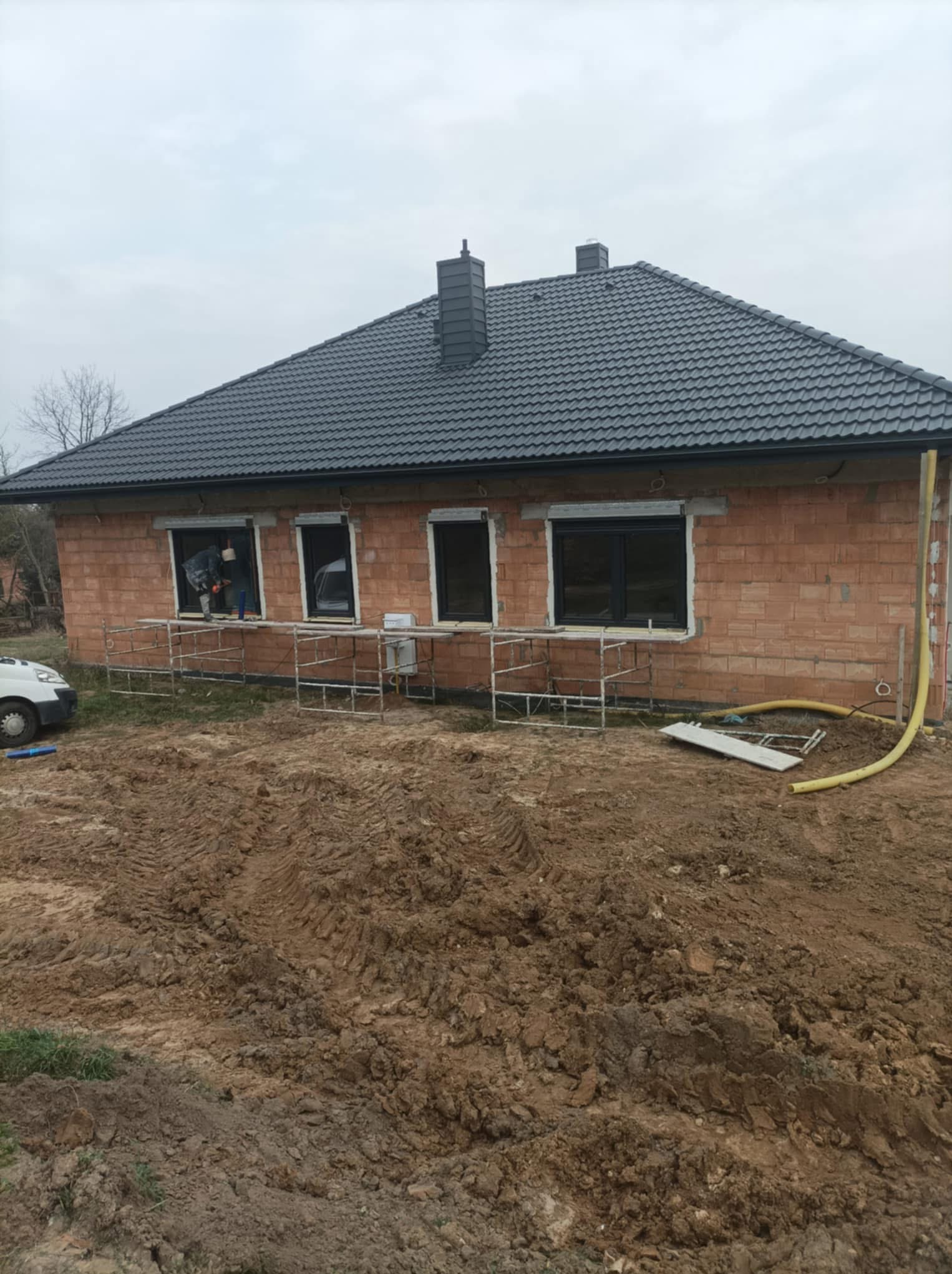 Dom w trakcie budowy z surowej cegły, widoczne rusztowania, okna bez wykończenia, przygotowany teren wokół budynku.