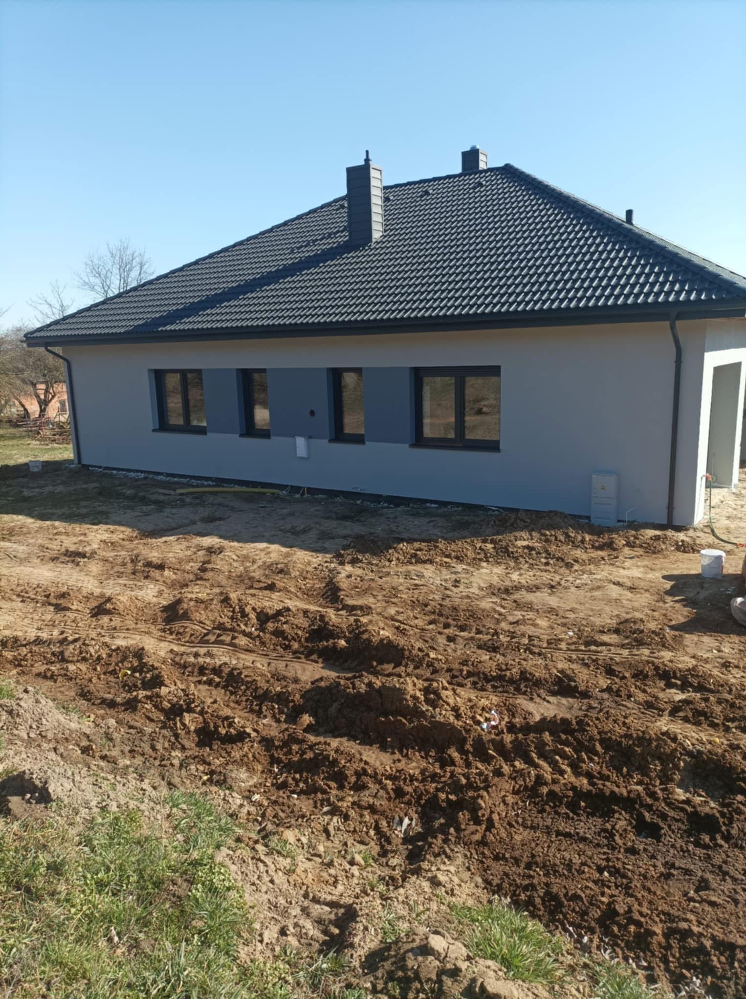 Nowoczesny dom jednorodzinny z szarą elewacją i grafitowym dachem, częściowo ocieplony, z widocznym niezagospodarowanym terenem wokół.