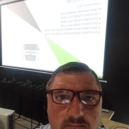 Klimatyzacja-Klima-Cool Jarek Pałka - Prezentacja klimatyzacji AURA w Gawłuszowicach. Mężczyzna w okularach na tle ekranu z modelami klimatyzatorów MULTISPLIT. Widoczny fragment klimatyzatora.