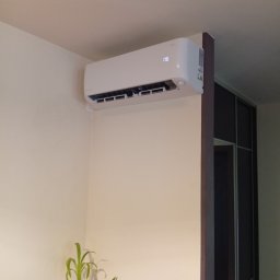 Klimatyzacja-Klima-Cool Jarek Pałka - Biały klimatyzator ścienny zamontowany w rogu pokoju, widoczny z dołu, z rośliną doniczkową w tle. Minimalistyczny design wnętrza.