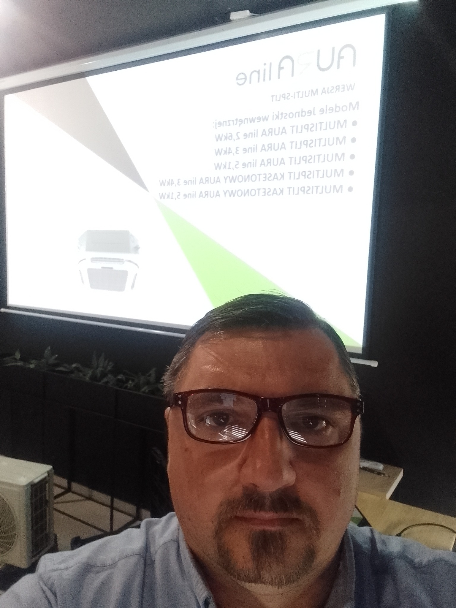 Prezentacja klimatyzacji AURA w Gawłuszowicach. Mężczyzna w okularach na tle ekranu z modelami klimatyzatorów MULTISPLIT. Widoczny fragment klimatyzatora.