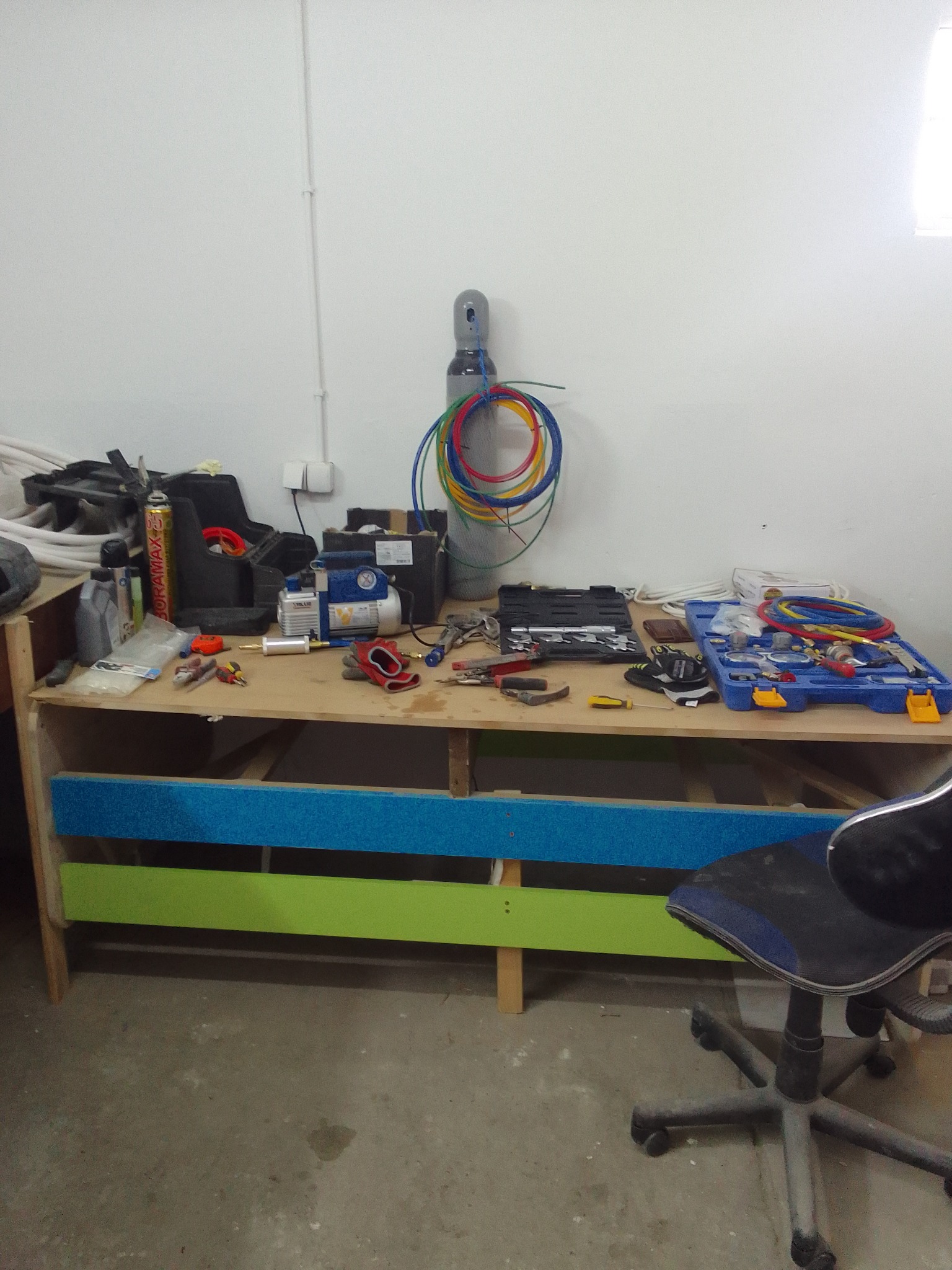 Warsztatowy stół z narzędziami i akcesoriami do klimatyzacji: butla z gazem, przewody, pompa próżniowa, skrzynka narzędziowa. Widok na stanowisko pracy serwisanta.