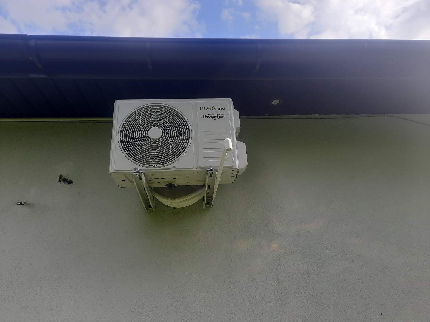 Biały klimatyzator Nura Line zamontowany na jasnej ścianie budynku, widoczny fragment dachu z rynną. Urządzenie w pełnej okazałości, z bliska, w naturalnym oświetleniu.