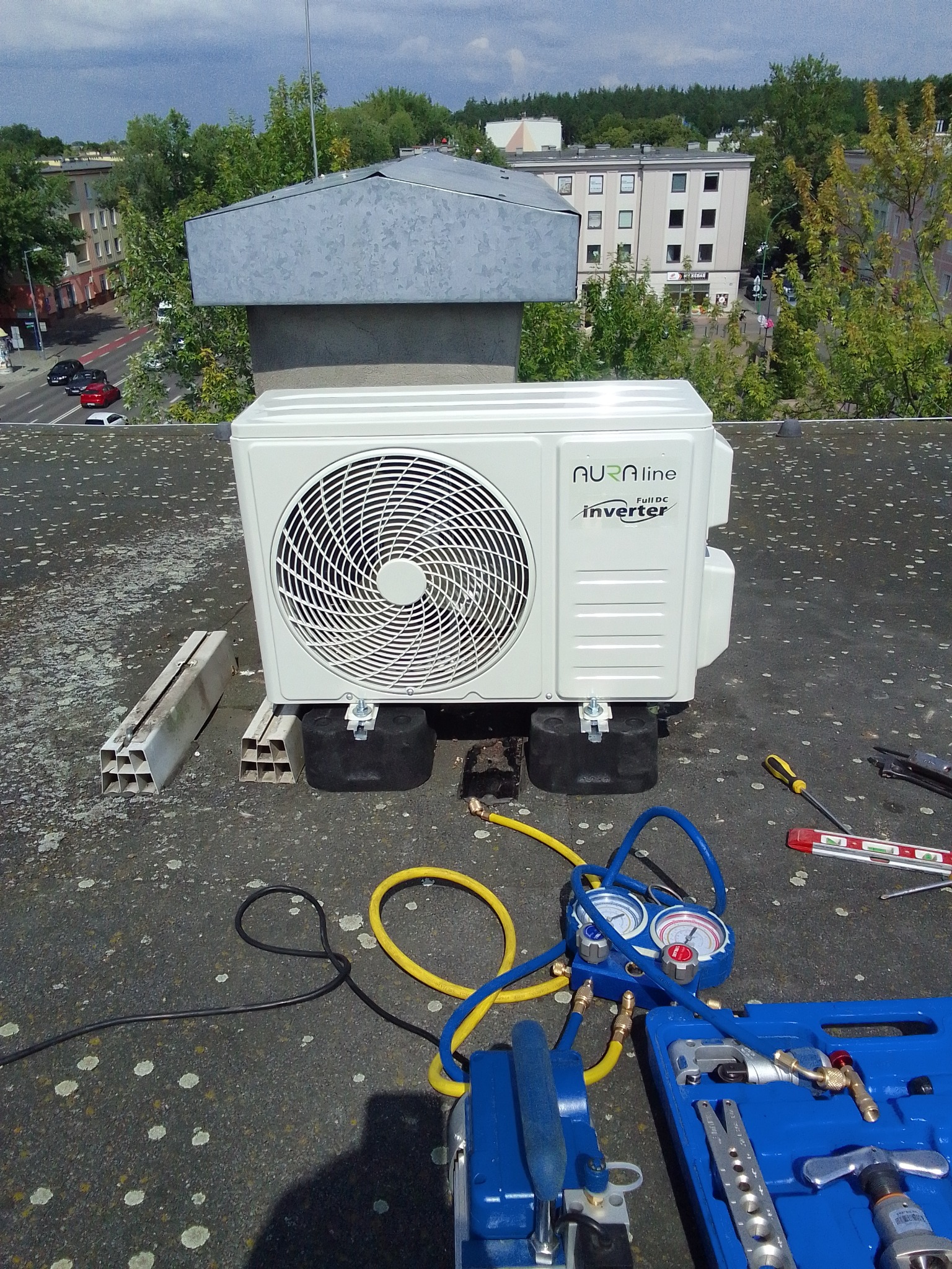 Biała jednostka zewnętrzna klimatyzacji Aura Line Full DC Inverter zamontowana na dachu budynku, widoczne narzędzia serwisowe i elementy montażowe.