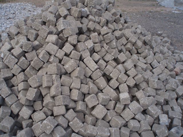 Stos szarych, surowych kostek o nieregularnych kształtach, ułożonych na ziemi. W tle widoczny fragment hałdy drobnego kruszywa.