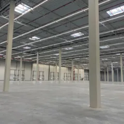 Wnętrze nowo wybudowanej, przestronnej hali z betonową posadzką, widoczne białe słupy nośne, szara konstrukcja dachu z oknami dachowymi i rzędami lamp, oraz segmentowe bramy wjazdowe po bokach.