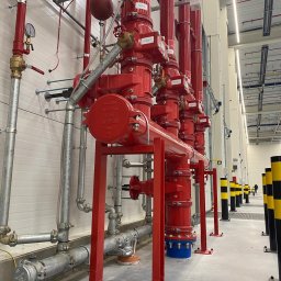 SpaceFactory CE Spółka z ograniczoną odpowiedzialnością spółka komandytowa - Czerwona instalacja przeciwpożarowa z rurami i zaworami w hali przemysłowej, widoczne metalowe podpory i czarno-żółte słupki odgradzające.