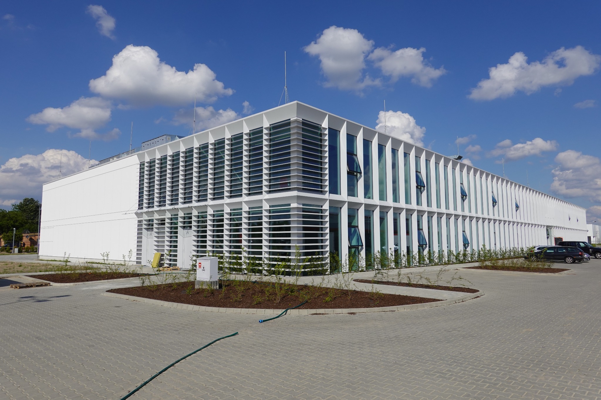 Nowoczesna, biała hala z licznymi oknami i żaluzjami, widok z zewnątrz, błękitne niebo z chmurami w tle, parking z kilkoma samochodami, świeżo posadzone krzewy wzdłuż budynku.
