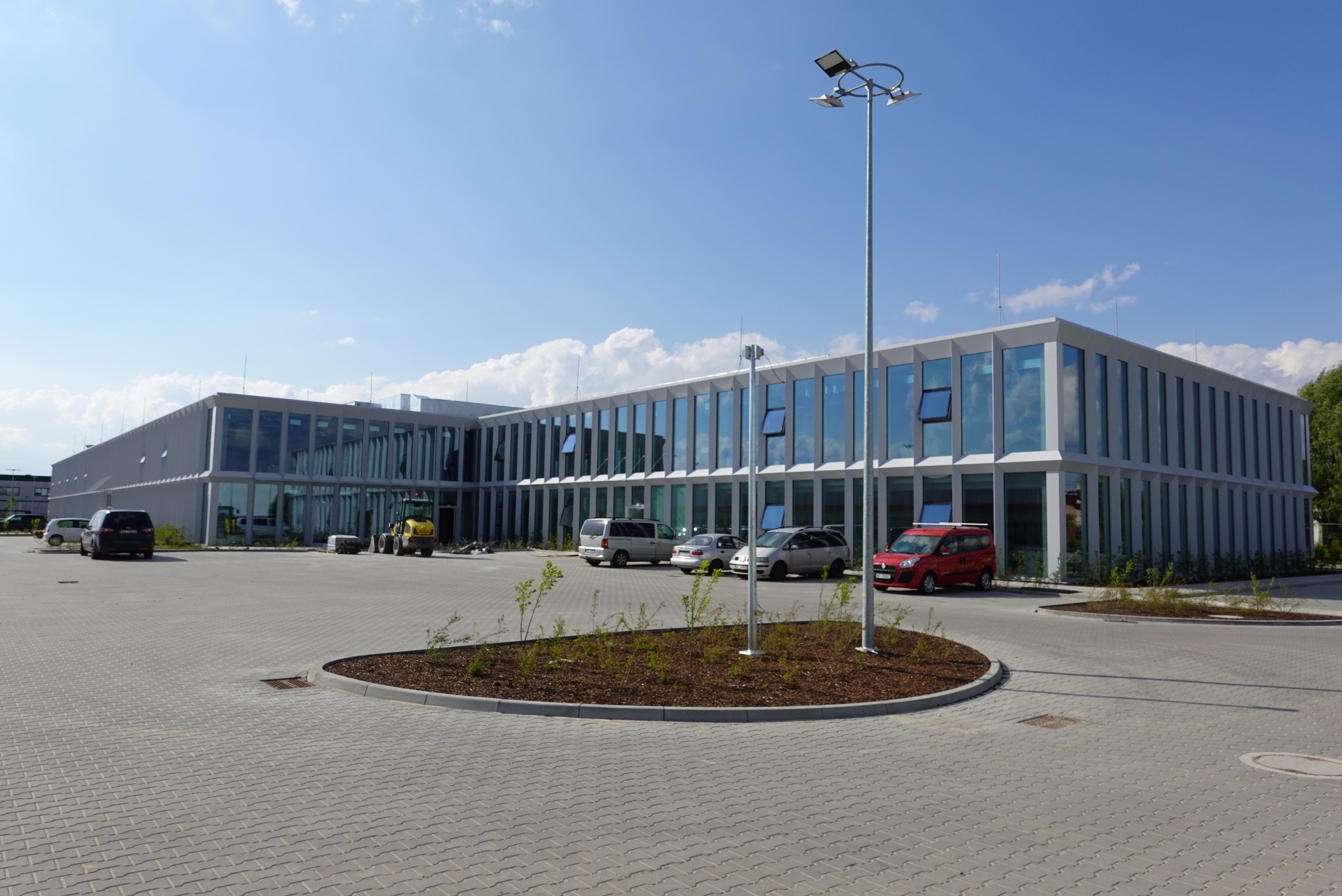 Nowoczesny budynek biurowy z dużą ilością przeszkleń, widoczny parking wyłożony kostką brukową i pojazdy, w tym mała koparka. Częściowo otwarte okna w budynku.