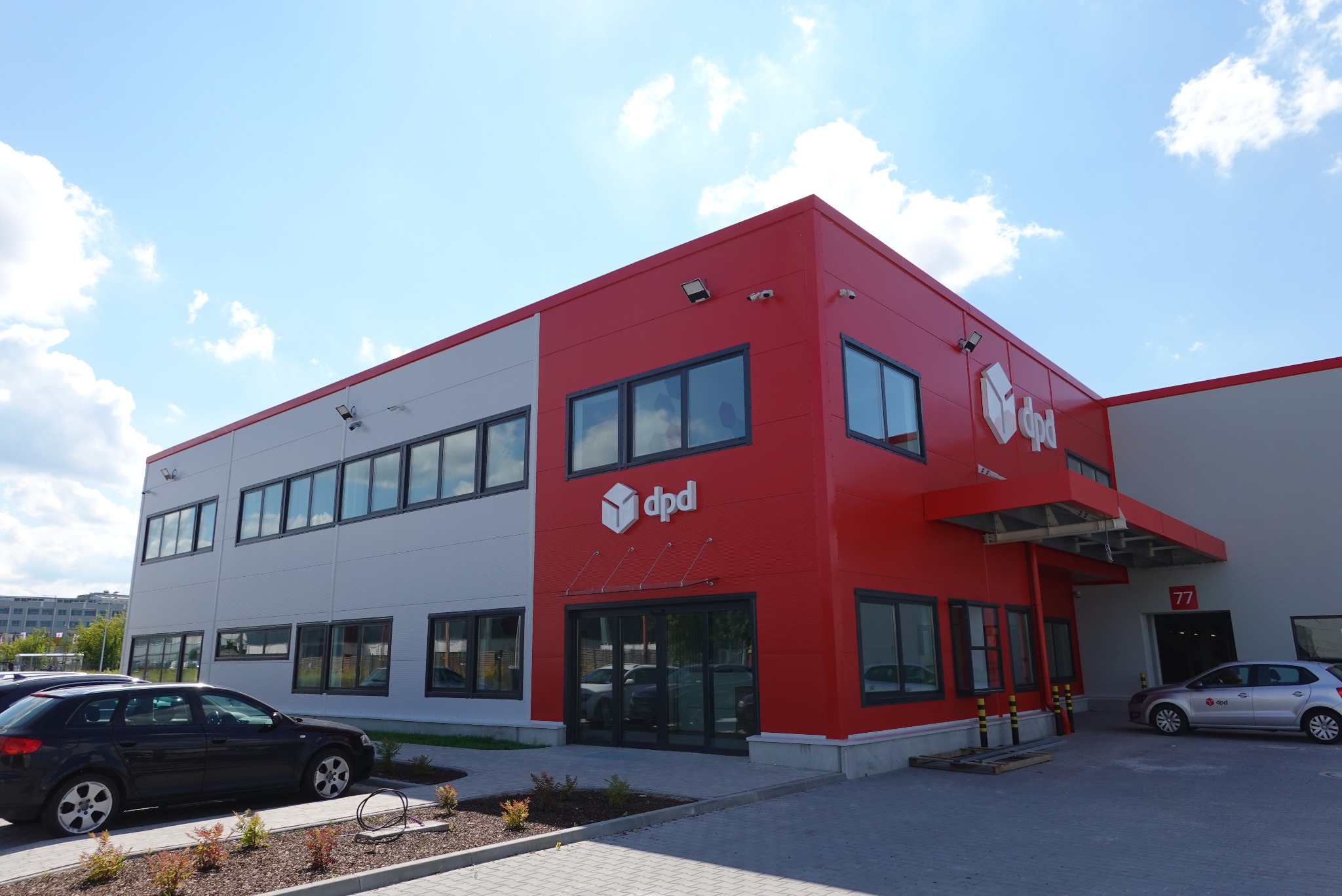 Nowoczesna hala magazynowa firmy kurierskiej DPD w Warszawie, z charakterystyczną czerwoną elewacją i logo, widoczna z zewnątrz w słoneczny dzień.