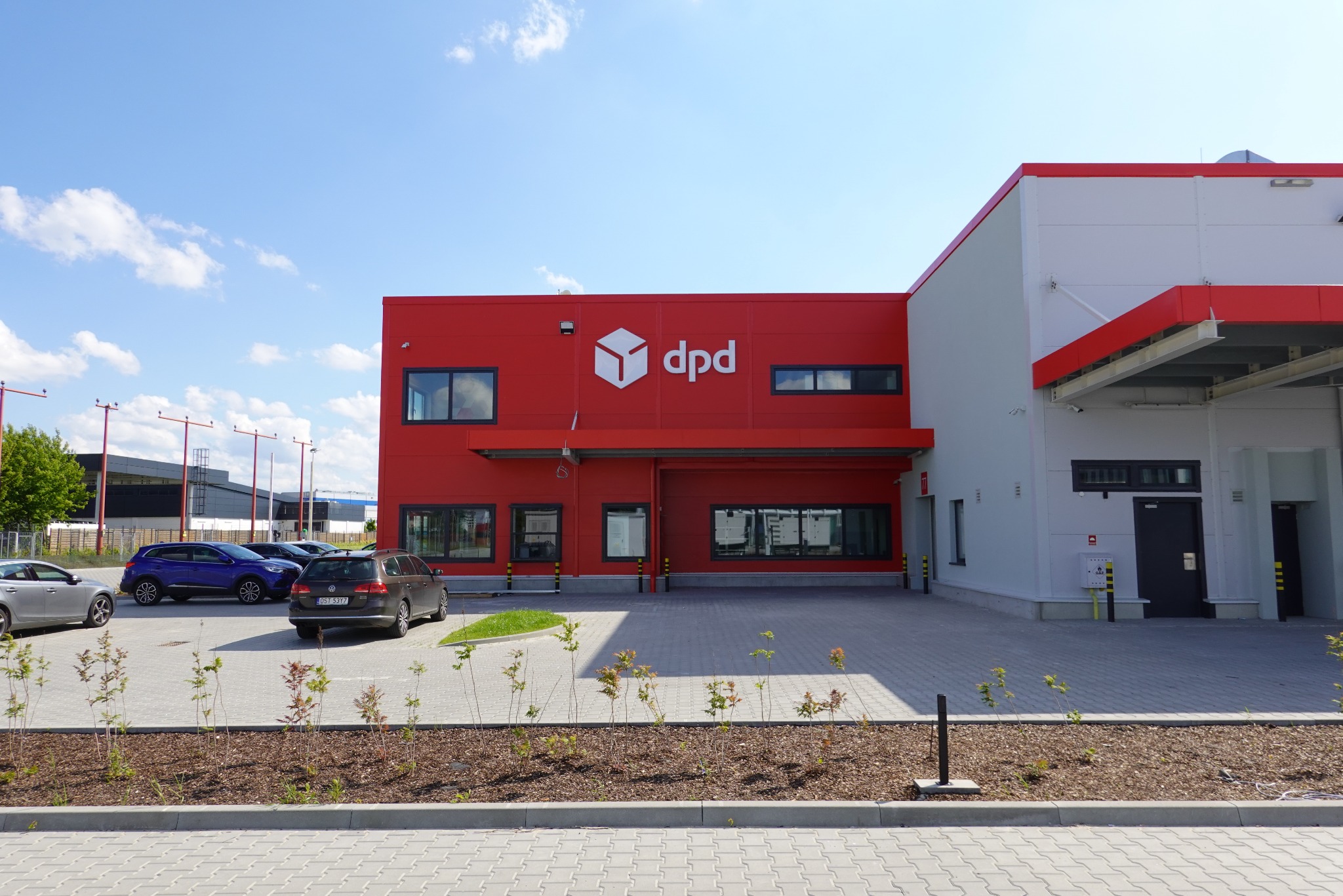 Czerwono-biała hala magazynowa z logo DPD, widok z zewnątrz, na pierwszym planie zaparkowane samochody i rabata z młodymi drzewkami.