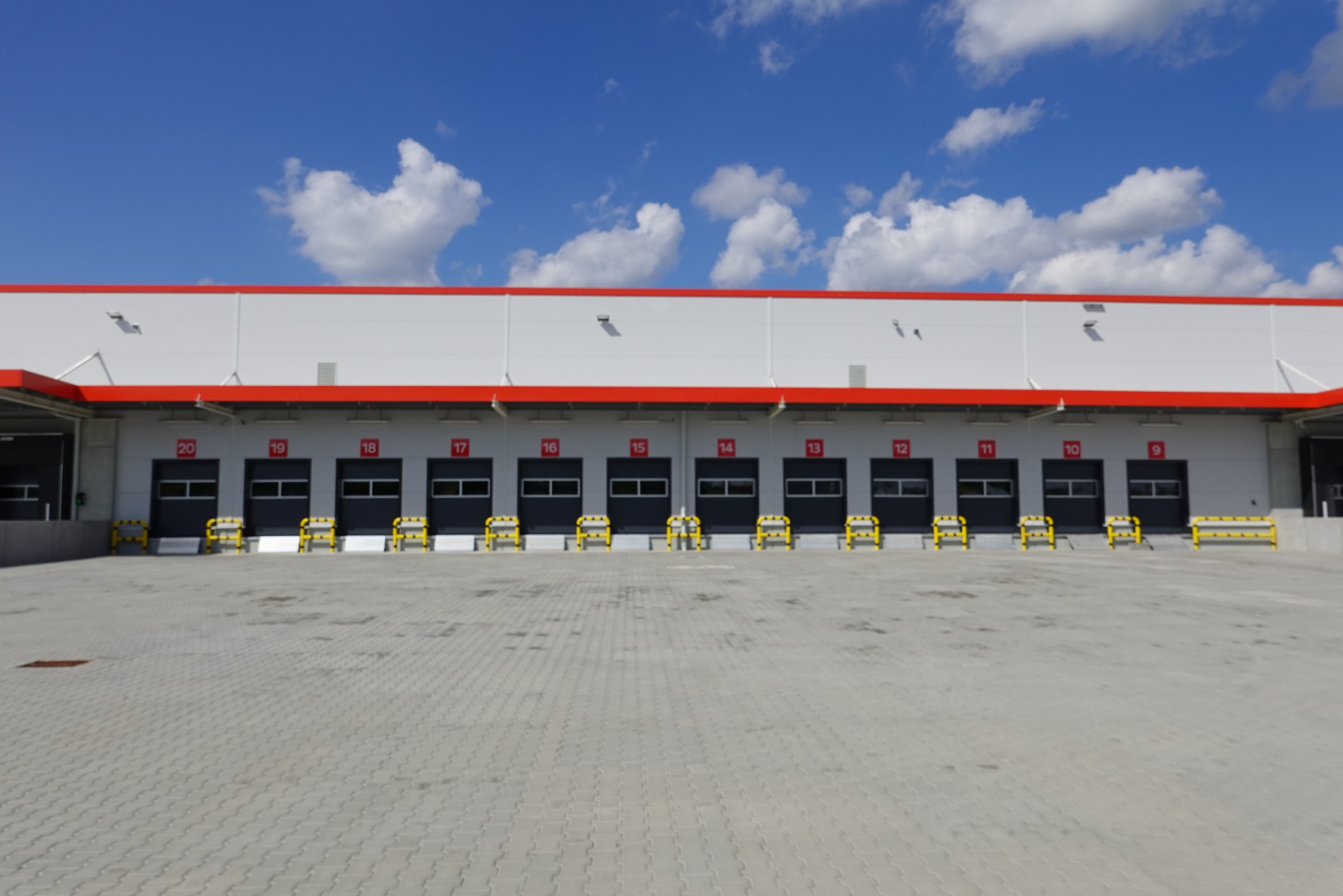 Fasada nowoczesnej hali magazynowej z ponumerowanymi bramami załadunkowymi, zabezpieczonymi żółtymi odbojami, na tle błękitnego nieba z obłokami.