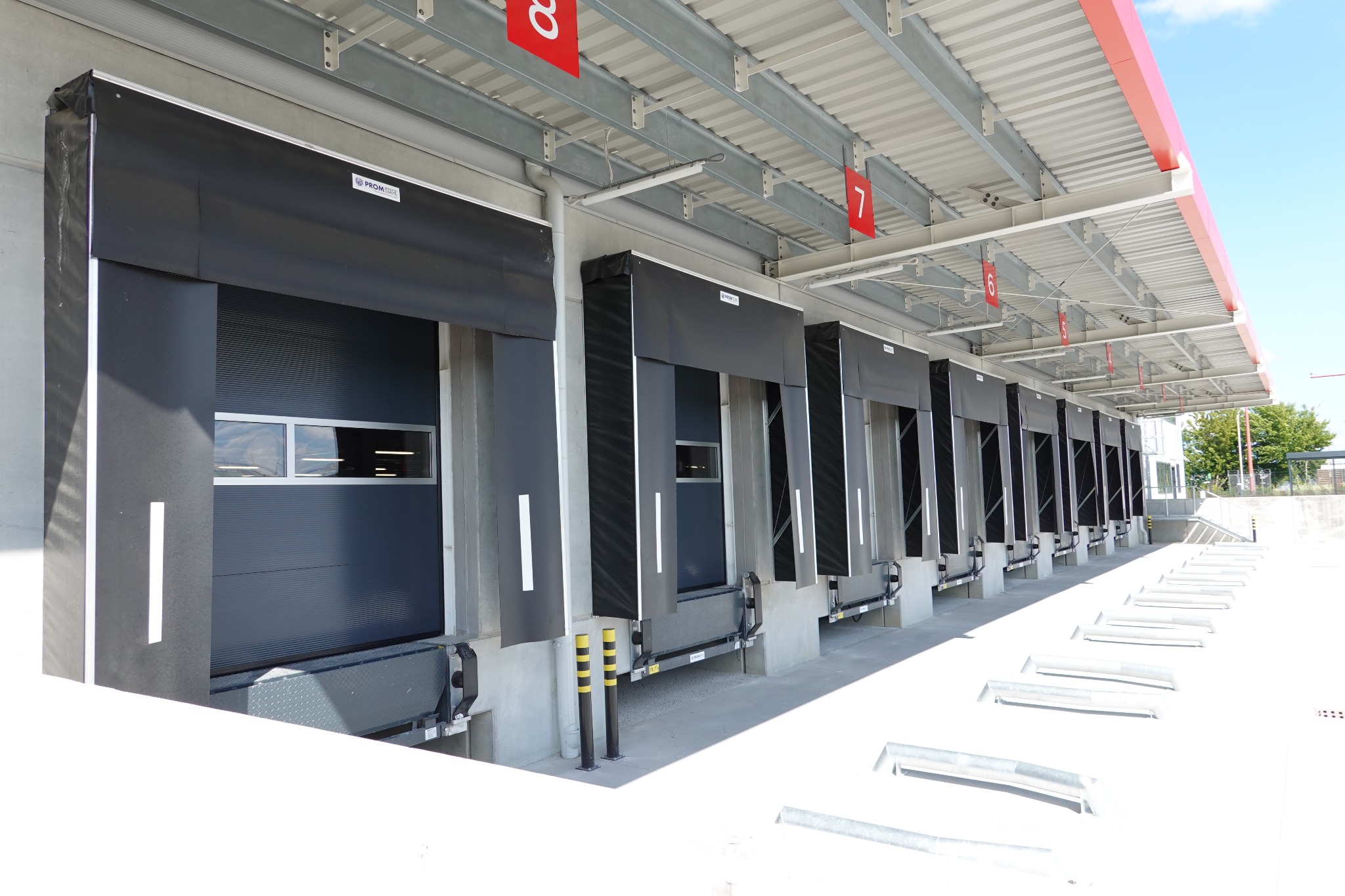 Rampa rozładunkowa hali magazynowej z czarnymi bramami segmentowymi, widoczne numery stanowisk 7 i 8, stalowa konstrukcja dachu i betonowe elementy elewacji.