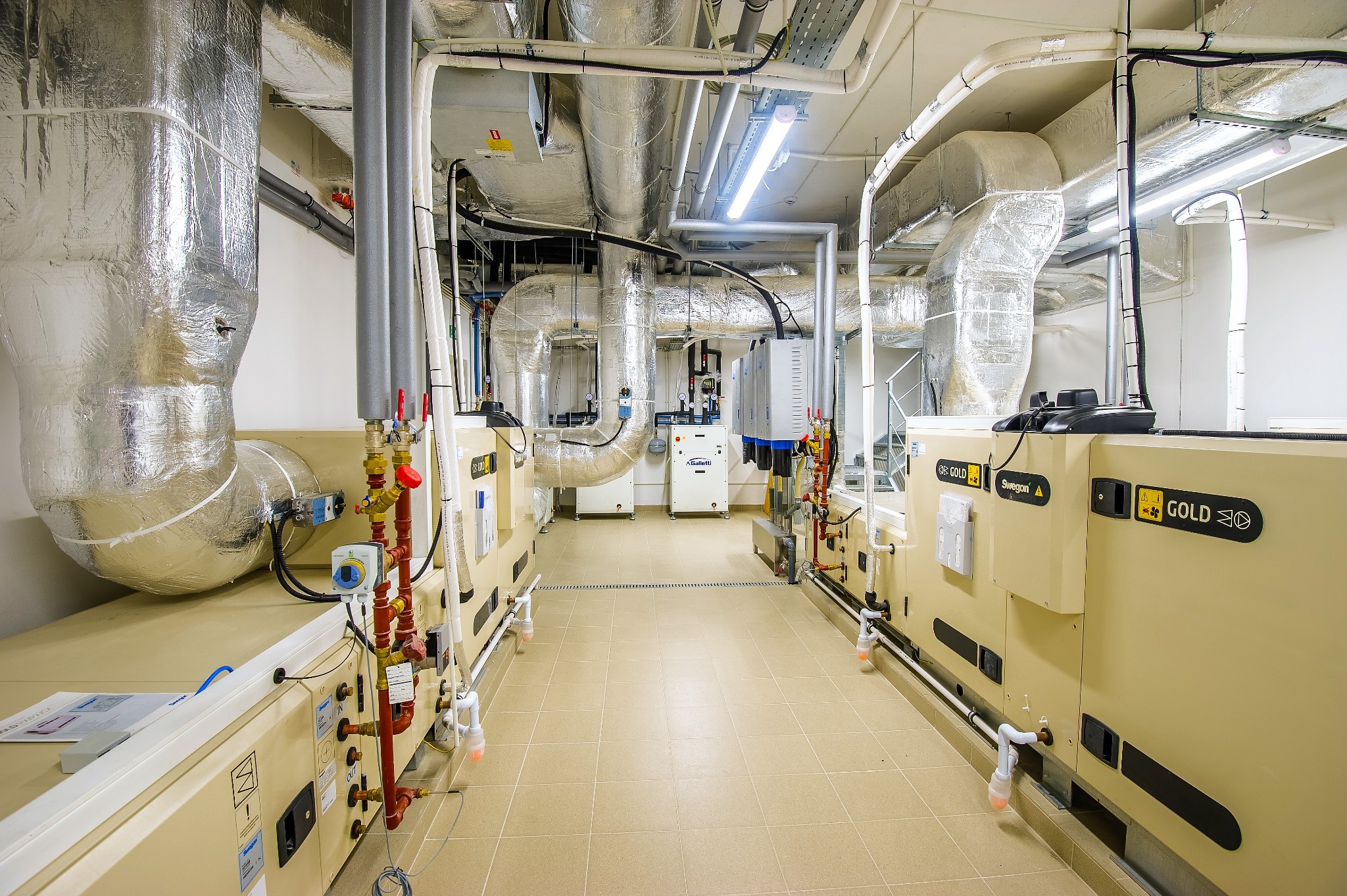 Wnętrze hali z systemem wentylacji i klimatyzacji, widoczne rury izolowane folią aluminiową, urządzenia Swegon Gold, instalacje rurowe z zaworami i manometrami, oświetlenie jarzeniowe.
