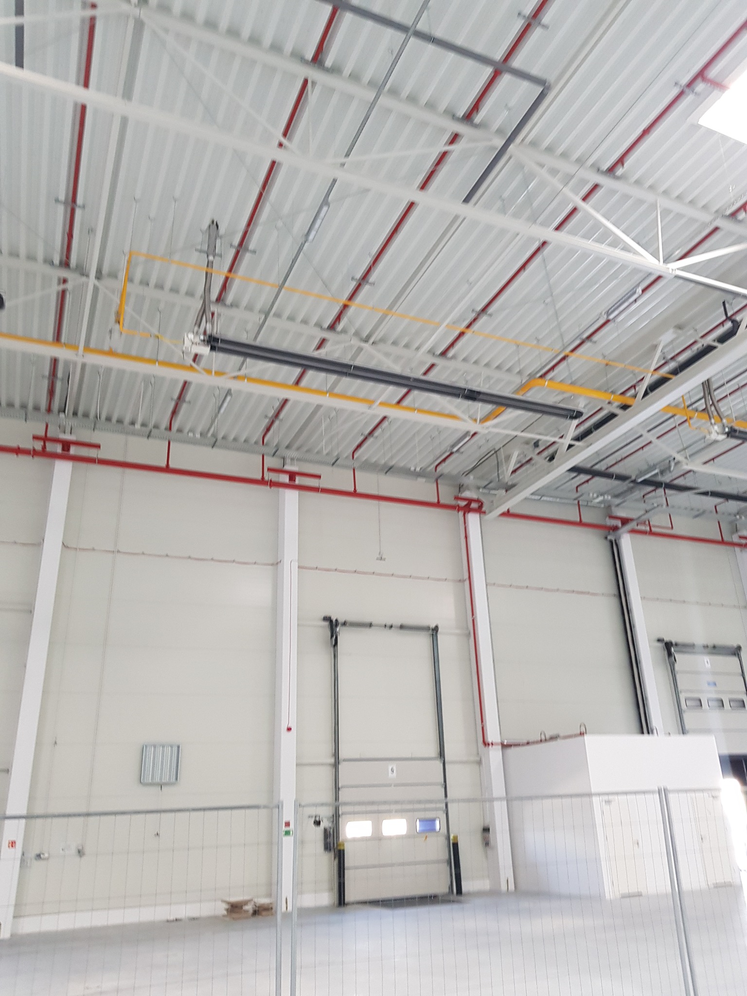 Wnętrze hali przemysłowej z widocznymi elementami konstrukcji stalowej dachu, instalacjami pod stropem oznaczonymi kolorami oraz bramami wjazdowymi.