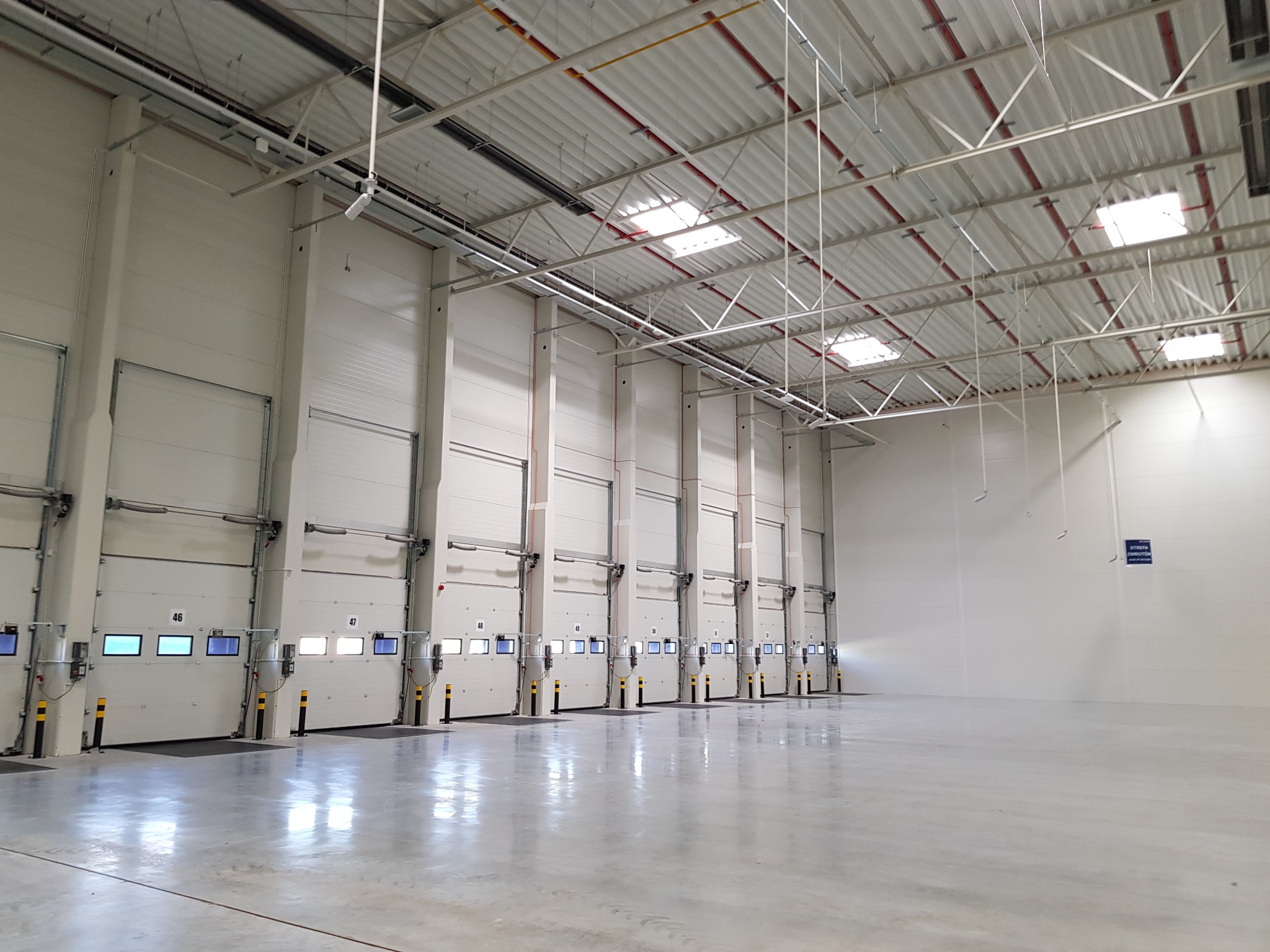 Wnętrze nowej, pustej hali magazynowej z szeregiem bram segmentowych z numerami, oświetlonych świetlikami dachowymi i lampami LED.