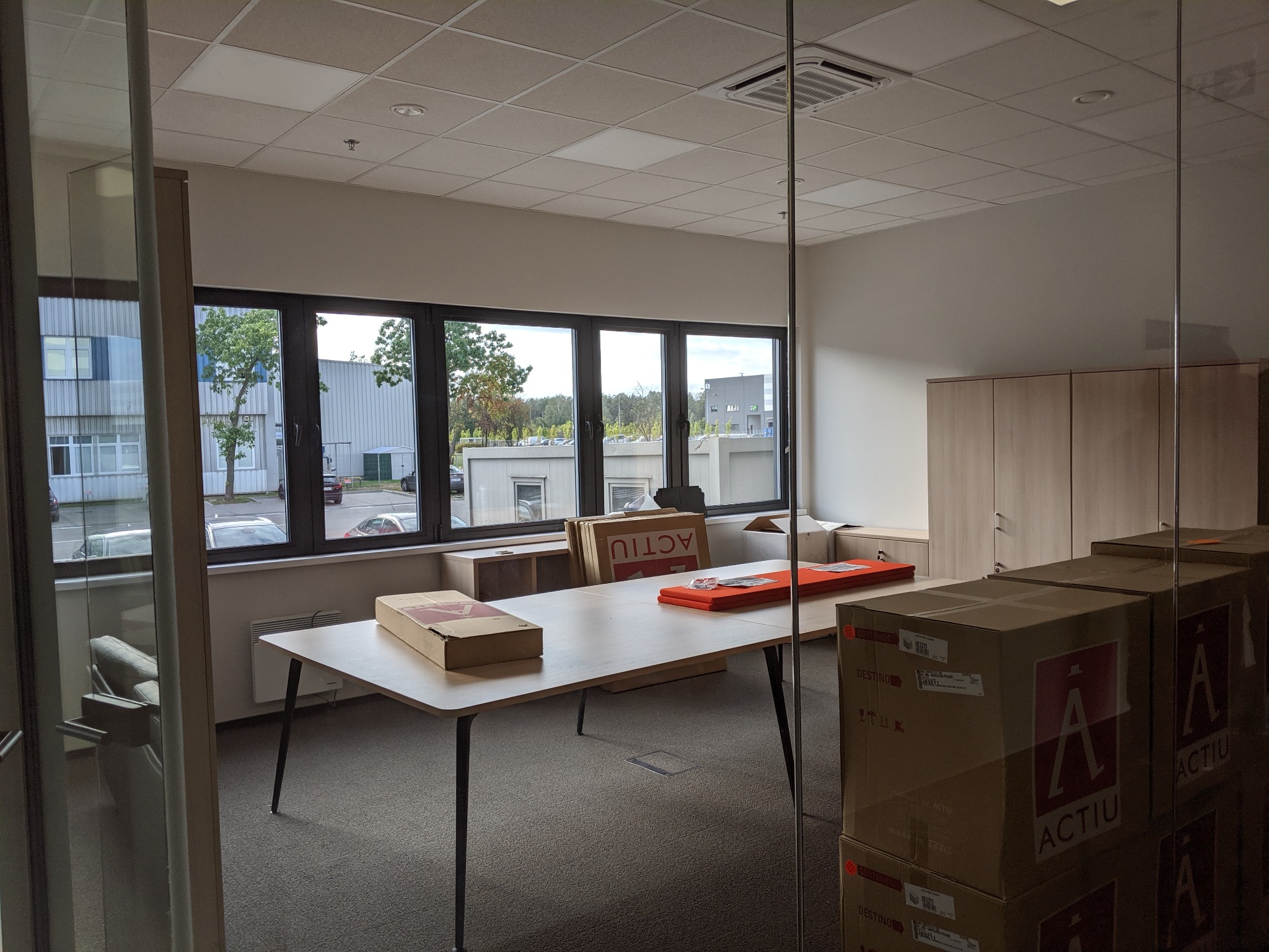 Wnętrze biura w hali produkcyjnej, widok przez szklaną ścianę, biurka z kartonami z logo ACTIU, okna z widokiem na zewnątrz, szafy w tle.