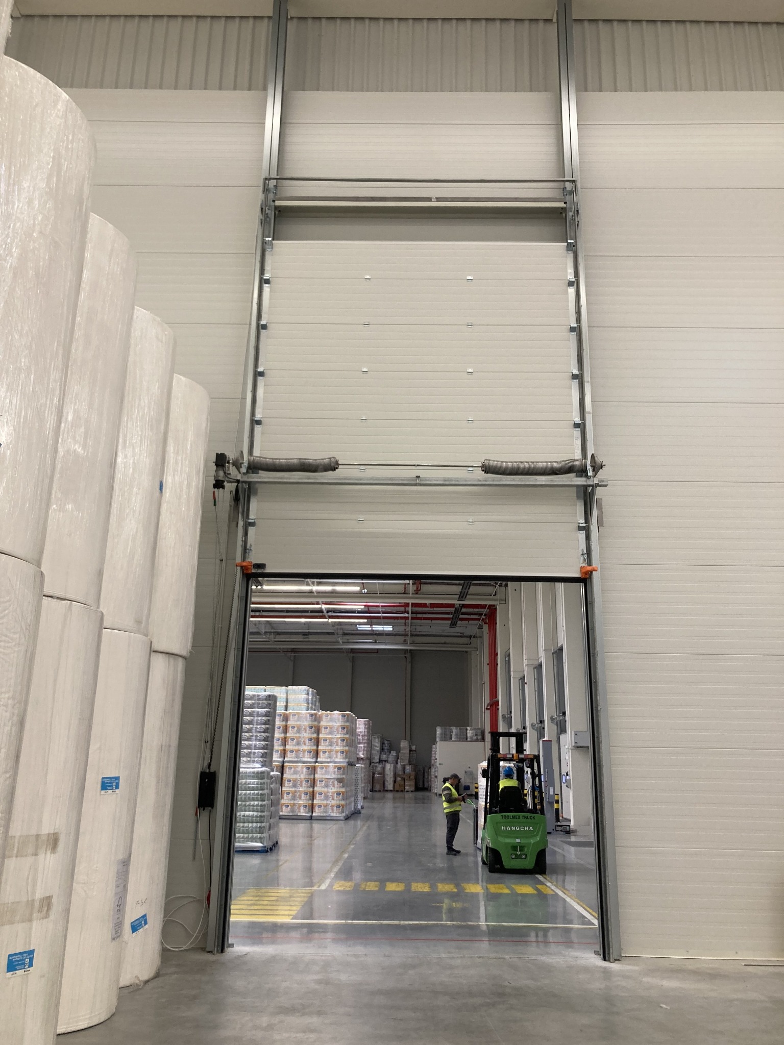 Wnętrze hali magazynowej z otwartą bramą segmentową; w tle widoczny wózek widłowy z operatorem w kamizelce odblaskowej i palety z towarem; po lewej stronie składowane rolki materiału.