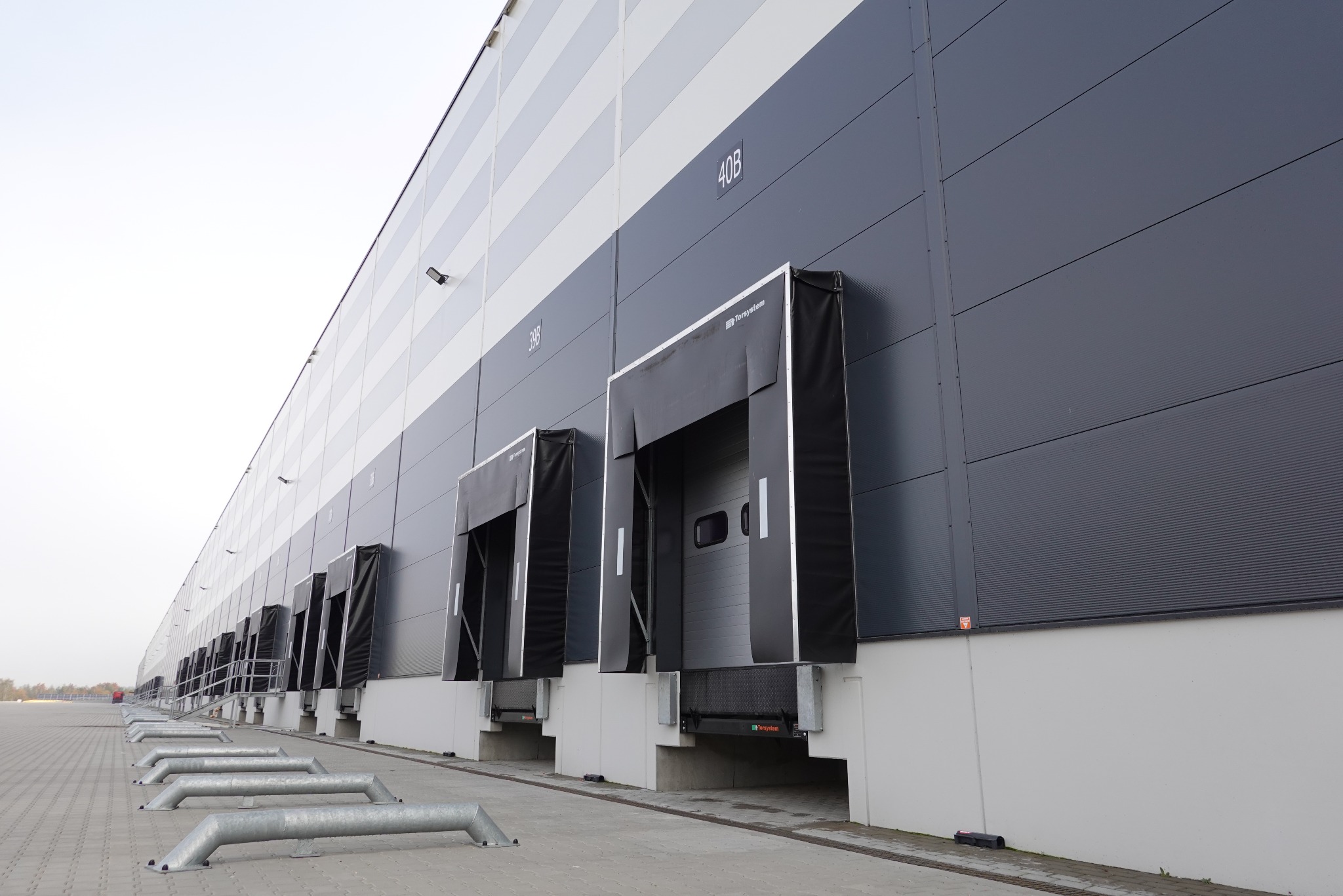 Długi budynek hali magazynowej z licznymi rampami załadunkowymi, zabezpieczonymi metalowymi barierami przed wjazdem samochodów.