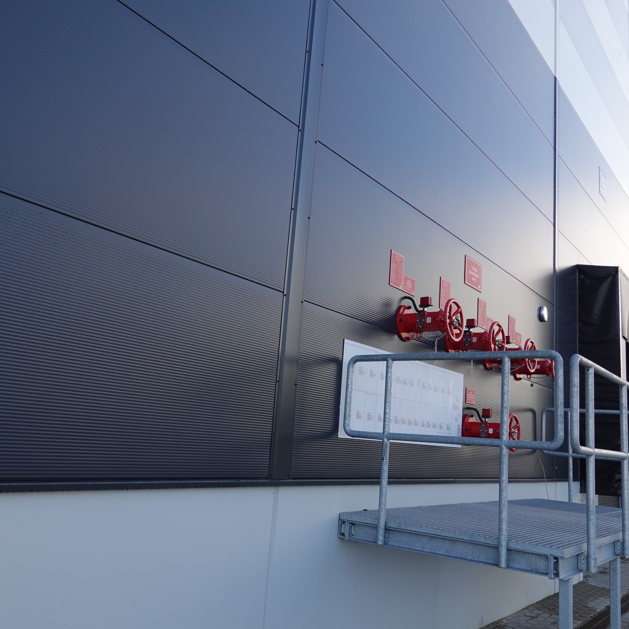 Zewnętrzna ściana hali z szarej blachy trapezowej z widocznymi zaworami przeciwpożarowymi w kolorze czerwonym i metalową platformą dostępową.