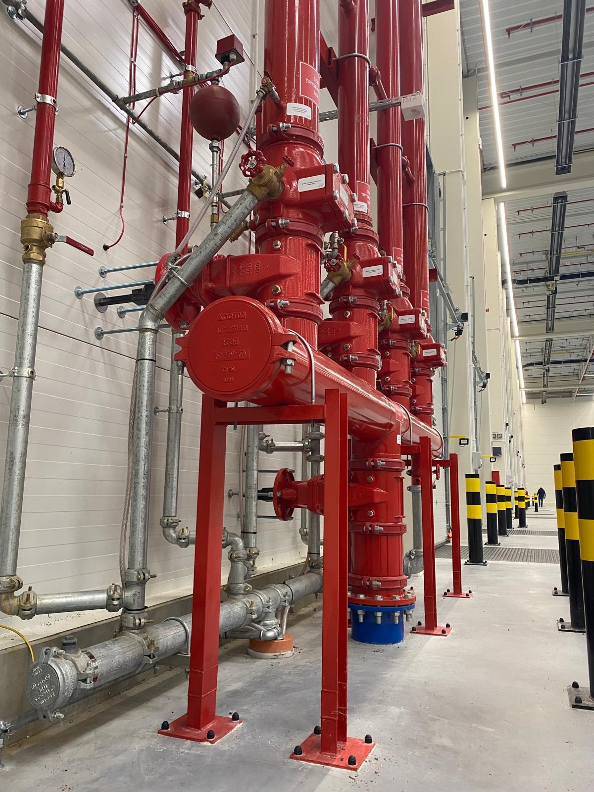 Czerwona instalacja przeciwpożarowa z rurami i zaworami w hali przemysłowej, widoczne metalowe podpory i czarno-żółte słupki odgradzające.