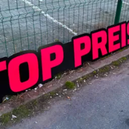 Tablica reklamowa z napisem 'TOP PREIS' w kolorze różowym, umieszczona przy chodniku. Kątowe ujęcie, widoczna siatka ogrodzeniowa w tle.