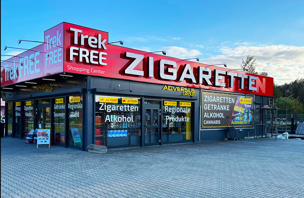 Fasada sklepu Trek Free Shopping Center w Łęknicy z dużym napisem 'Zigaretten' i ofertą: napoje, alkohol, regionalne produkty. Widoczne reklamy i stojak KAS.