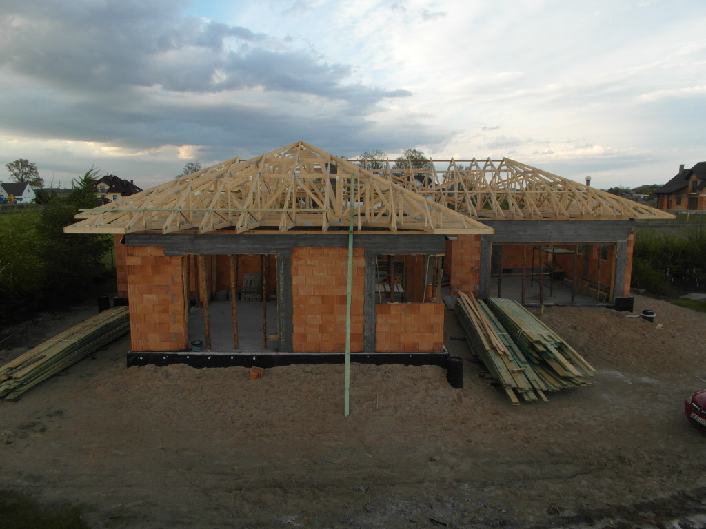 Budowa domu jednorodzinnego w trakcie wznoszenia ścian z cegły i konstrukcji drewnianej dachu, z widocznymi belkami i elementami konstrukcyjnymi.