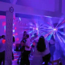 Tańczący ludzie w zadymionym pomieszczeniu oświetlonym stroboskopami i dekoracjami, w tle biała kolumna z naklejoną tabliczką '50'.