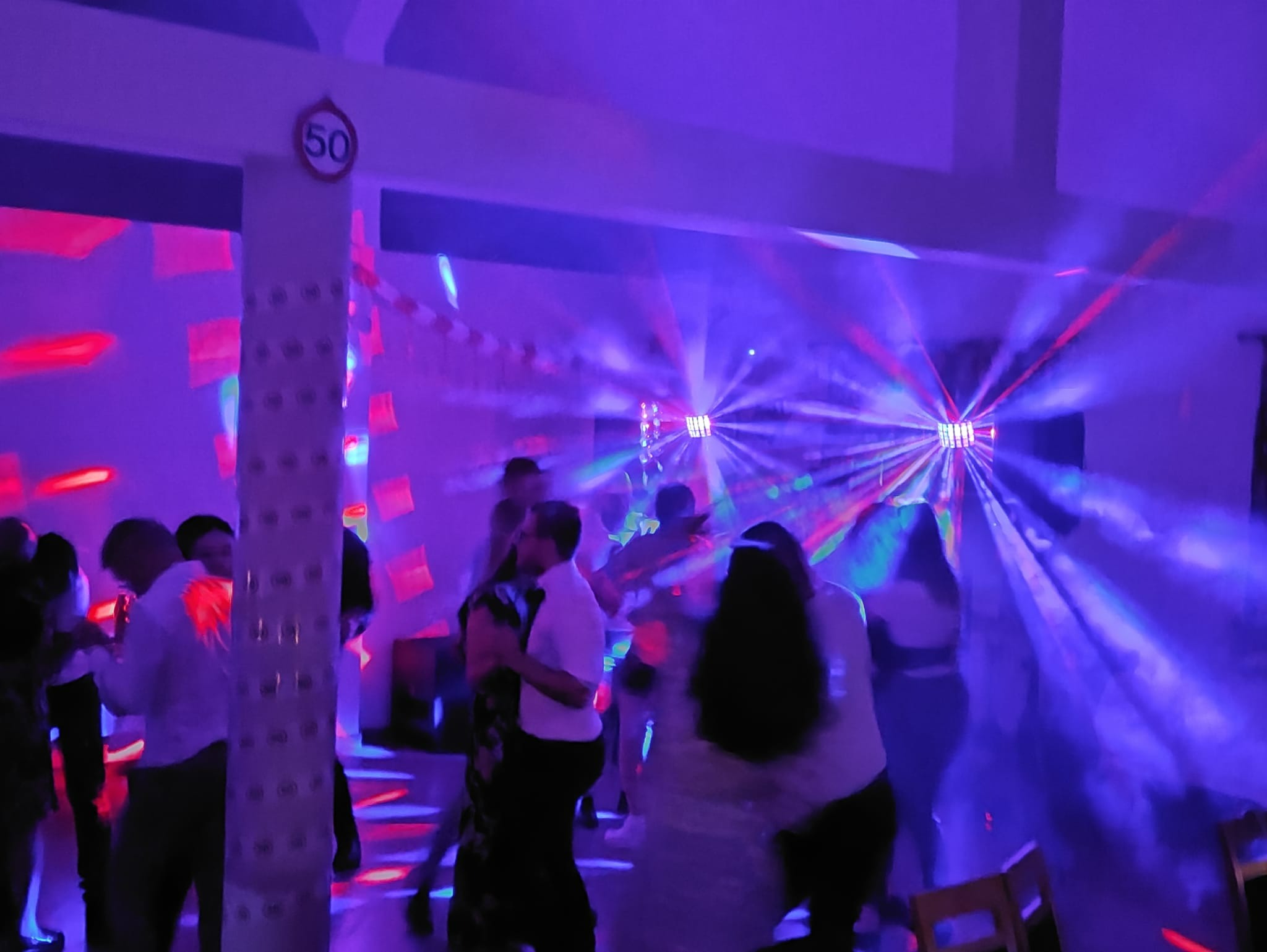 Tańczący ludzie w zadymionym pomieszczeniu oświetlonym stroboskopami i dekoracjami, w tle biała kolumna z naklejoną tabliczką '50'.