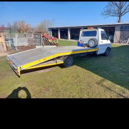 DAVKAS Dawid Łukacki - Biała laweta z żółtymi elementami na trawiastym terenie, z budynkiem gospodarczym w tle, w słoneczny dzień. Widoczny cień osoby robiącej zdjęcie.