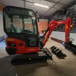 Czerwona mini koparka Kubota KX019-4 z łyżkami leży na betonowej posadzce magazynu, na tle palet z czarnymi skrzynkami i ściany z cegieł.