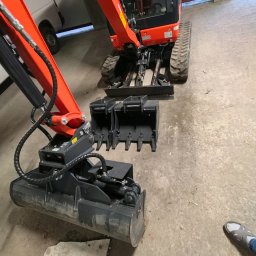 DAVKAS Dawid Łukacki - Pomarańczowa mini koparka z czarnym osprzętem na betonowej posadzce, widok z góry, częściowo widoczne stopy osoby w obuwiu. W tle biały samochód dostawczy.