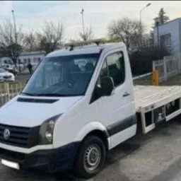 Biała laweta Volkswagen Crafter z platformą załadunkową na parkingu miejskim w pochmurny dzień. Widoczne detale pojazdu i otoczenie miejskie.