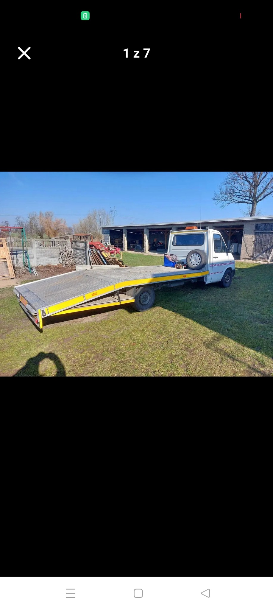 Biała laweta z żółtymi elementami na trawiastym terenie, z budynkiem gospodarczym w tle, w słoneczny dzień. Widoczny cień osoby robiącej zdjęcie.