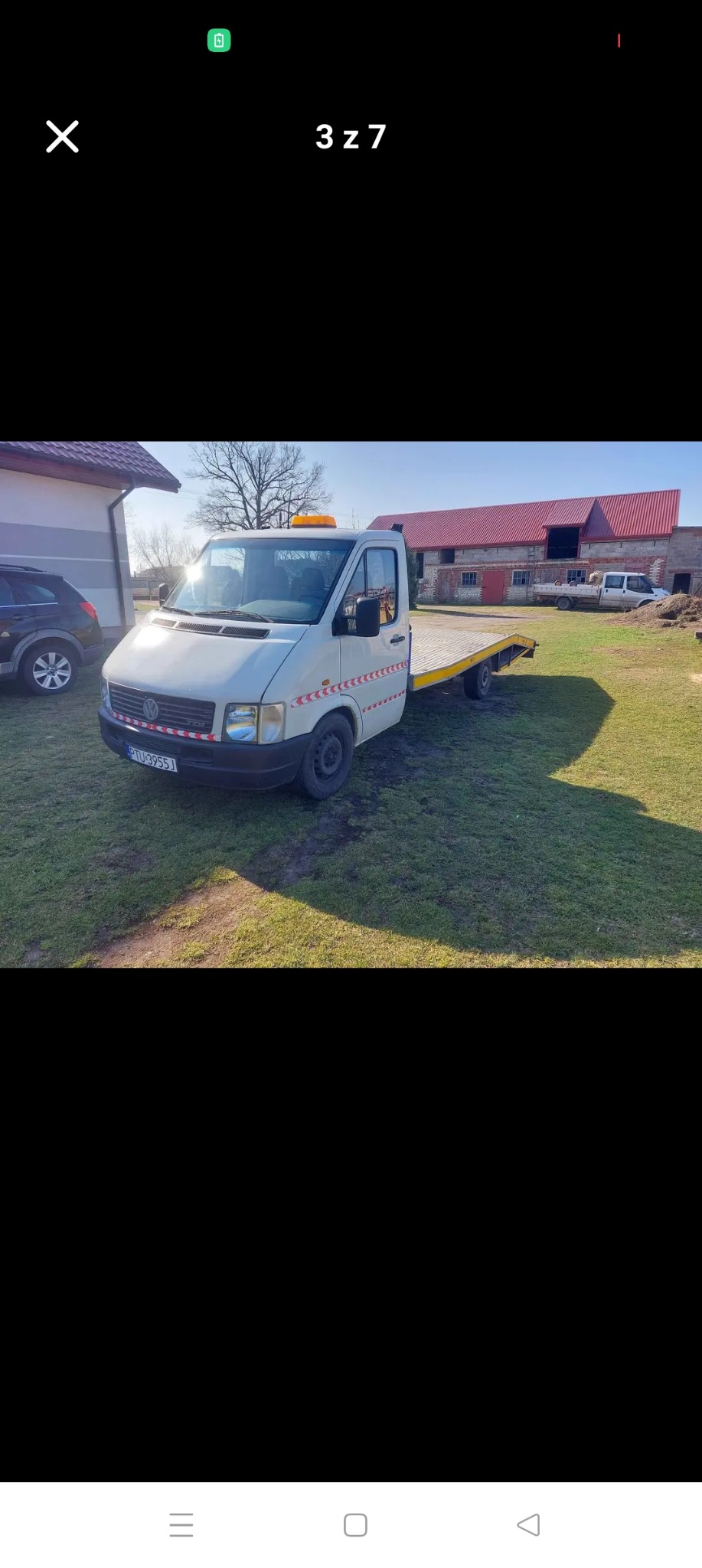 Biała laweta VW LT z pomarańczowym kogutem, zaparkowana na trawie przed budynkiem gospodarczym z czerwonym dachem. Widoczny fragment samochodu osobowego.