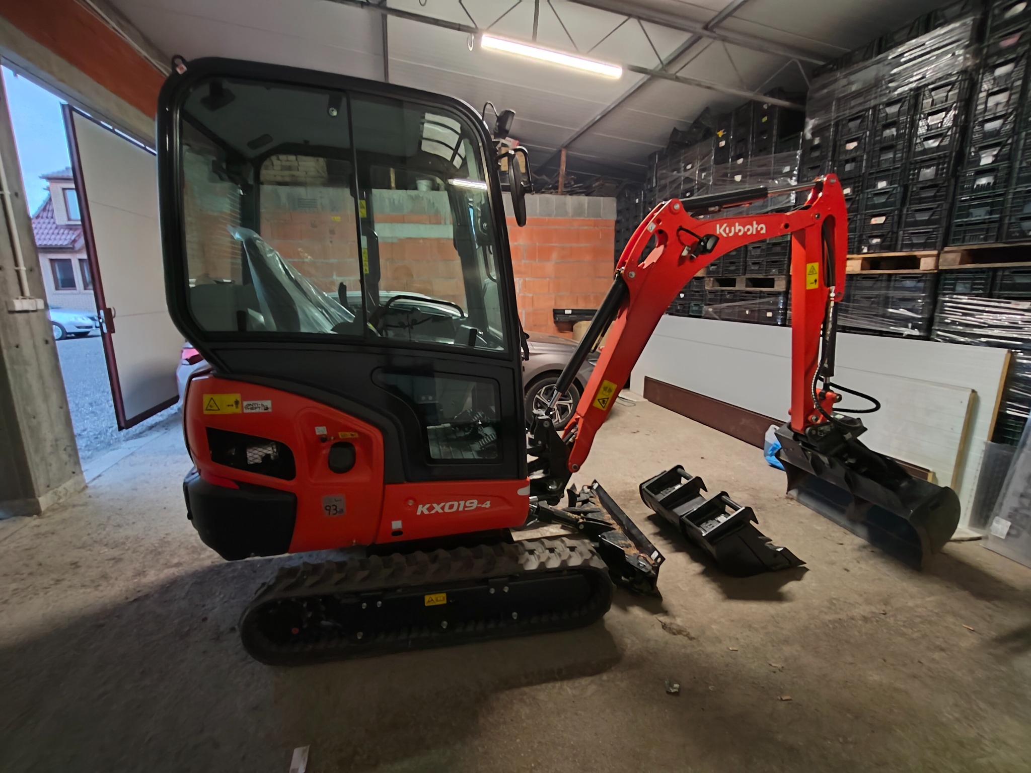 Czerwona mini koparka Kubota KX019-4 z łyżkami leży na betonowej posadzce magazynu, na tle palet z czarnymi skrzynkami i ściany z cegieł.