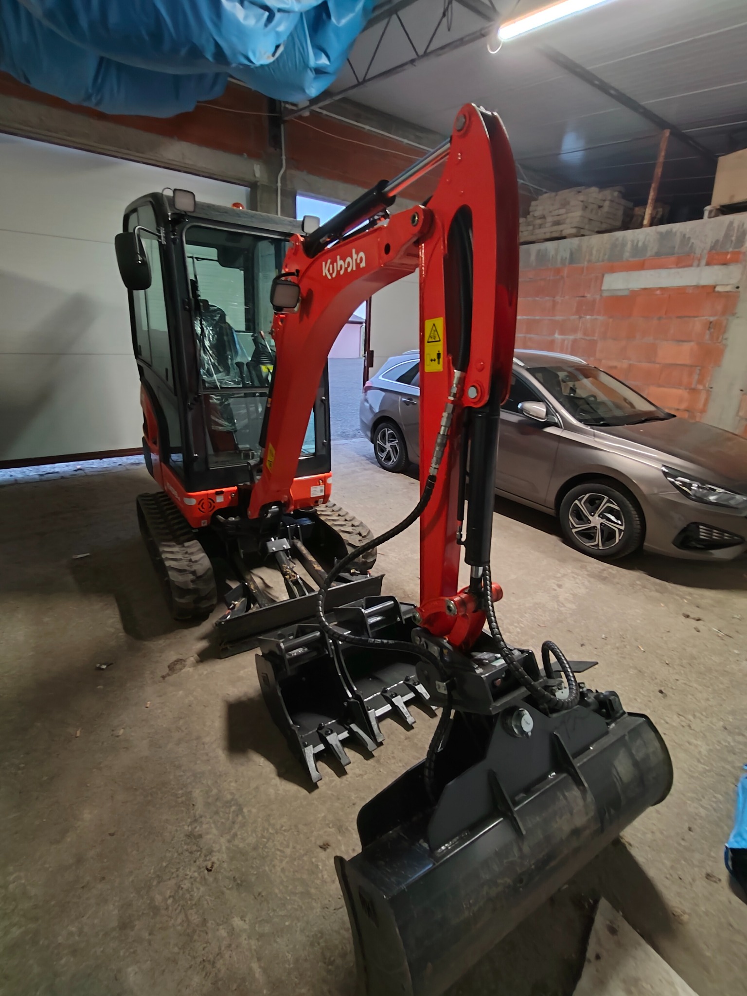 Czerwona mini koparka Kubota z czarną łyżką i osprzętem, stoi w garażu na betonowej posadzce. W tle szary samochód osobowy i ściana z cegły.