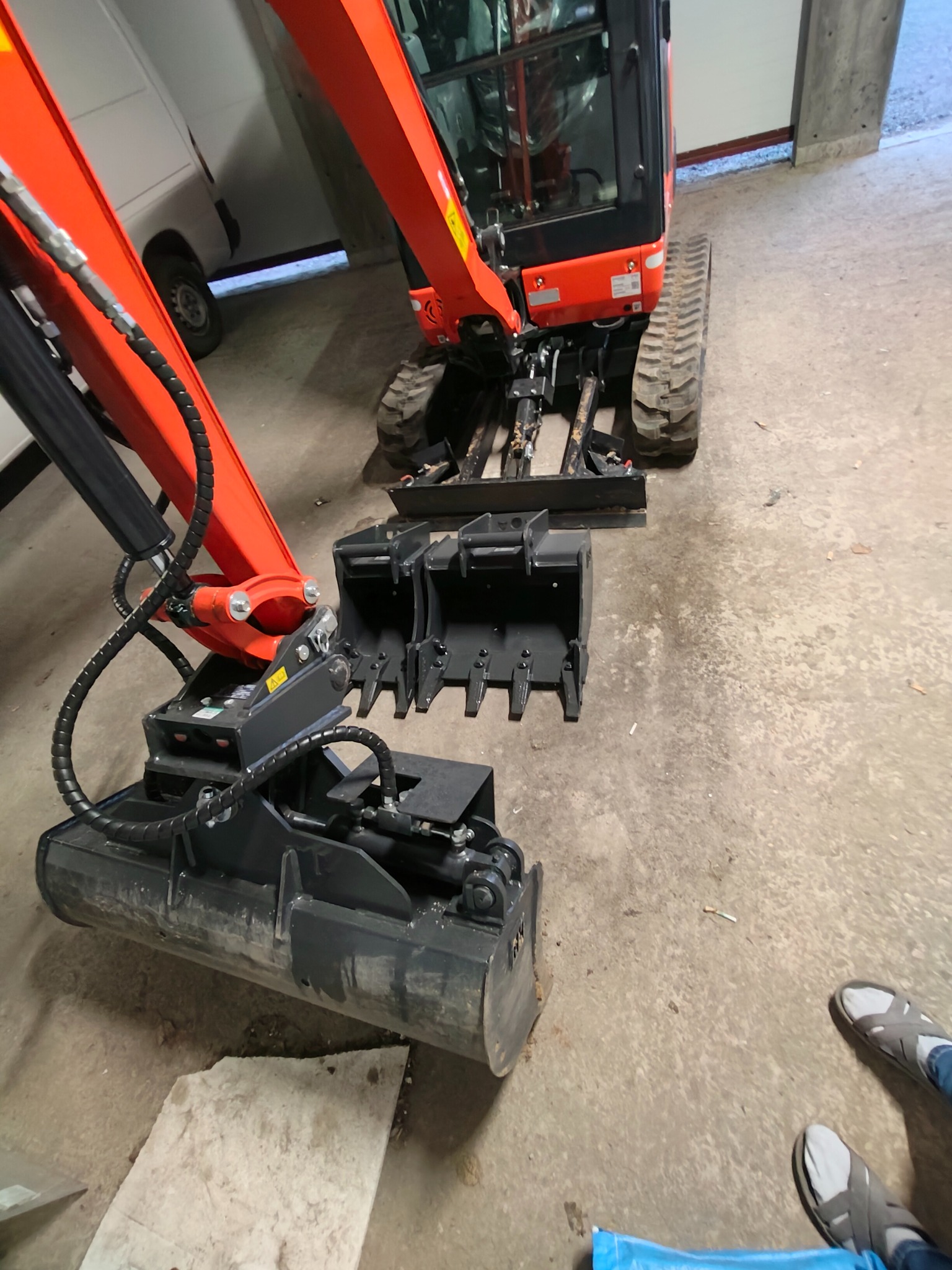 Pomarańczowa mini koparka z czarnym osprzętem na betonowej posadzce, widok z góry, częściowo widoczne stopy osoby w obuwiu. W tle biały samochód dostawczy.