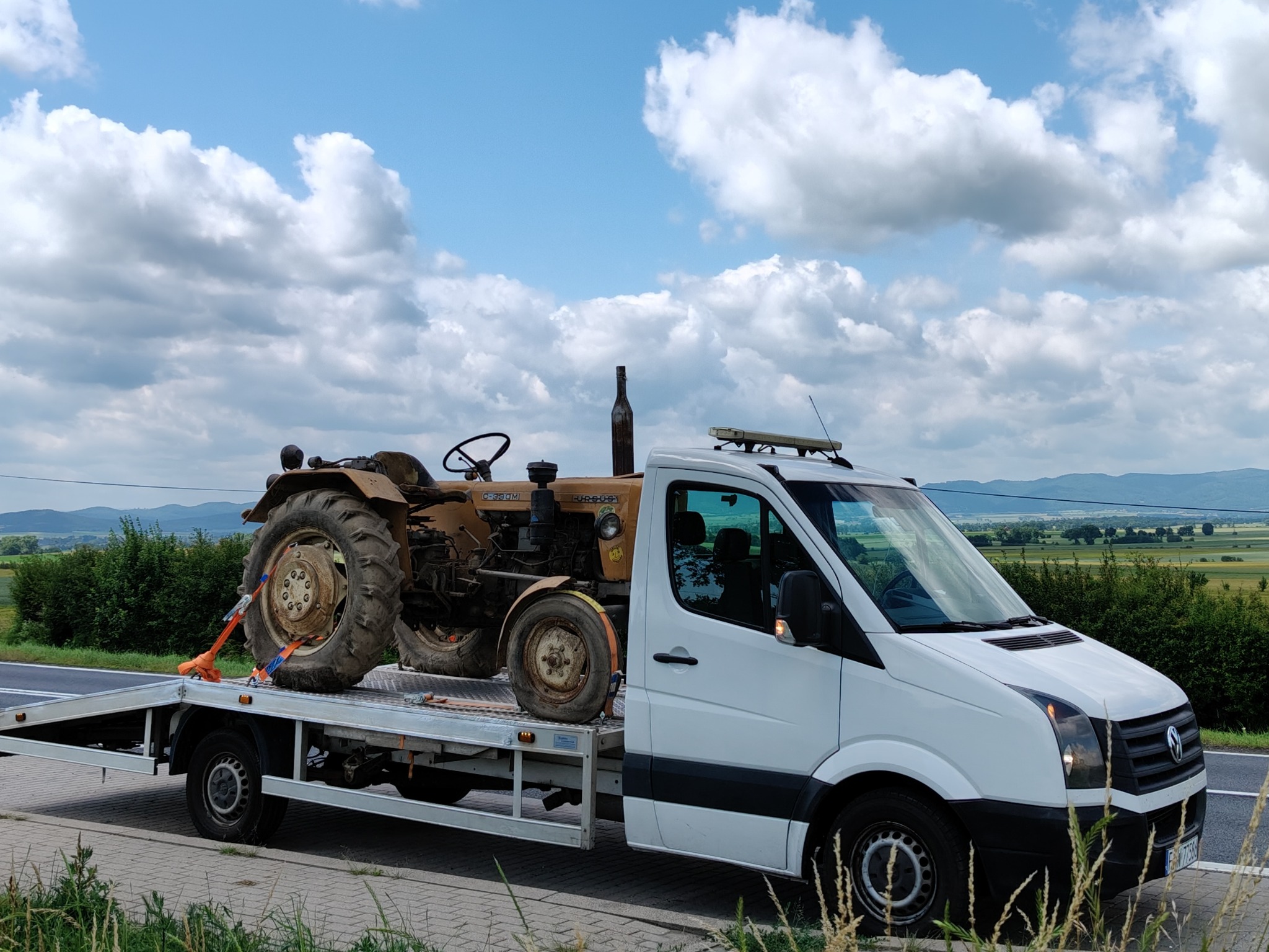 Biała laweta z traktorem Ursus C-330M zabezpieczonym pasami transportowymi na platformie. W tle błękitne niebo z obłokami i zielony krajobraz.