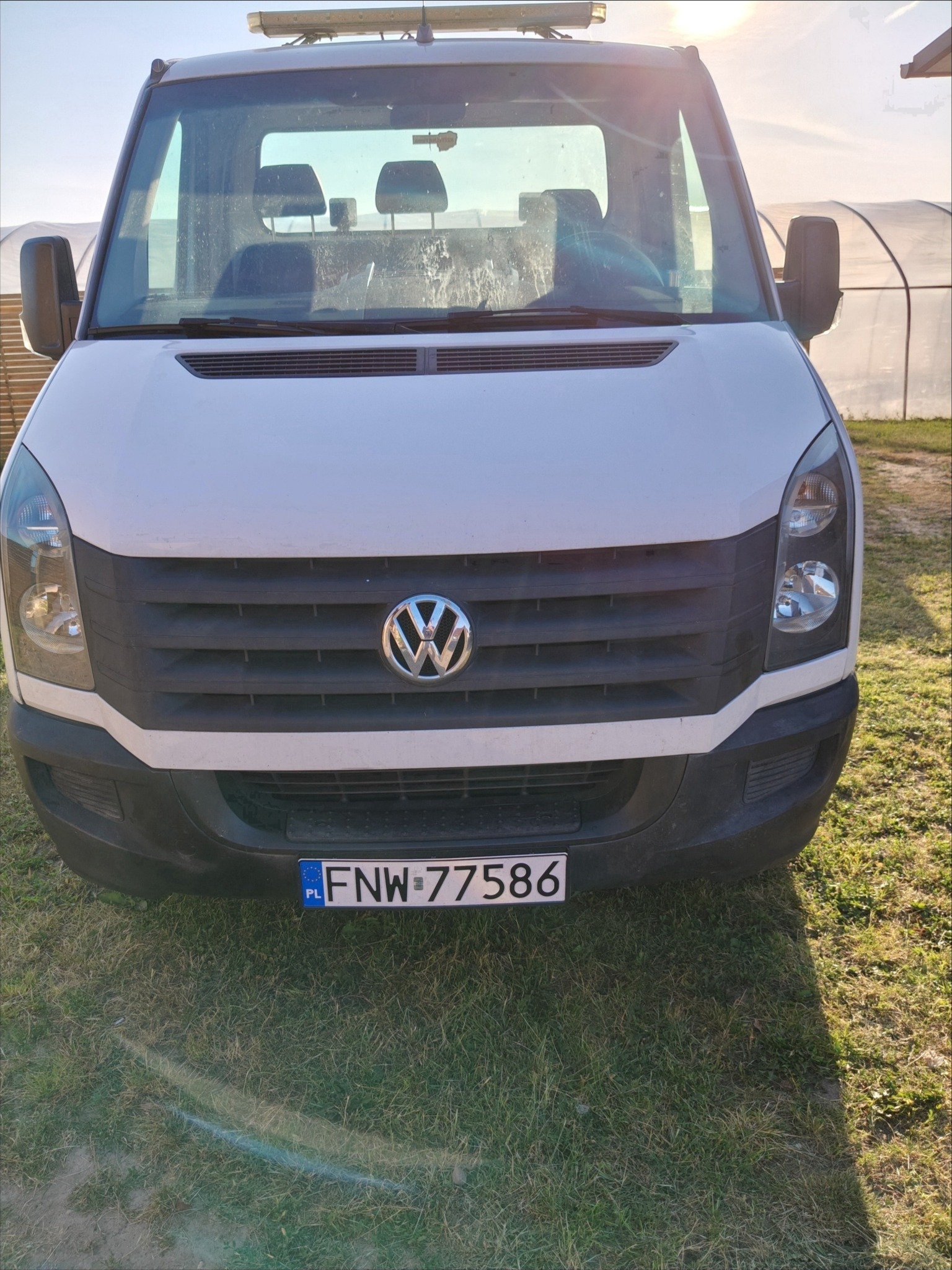 Biały Volkswagen Crafter z polską tablicą rejestracyjną FNW-77586, zaparkowany na trawie, z widocznym przodem pojazdu i fragmentem szklarni w tle.