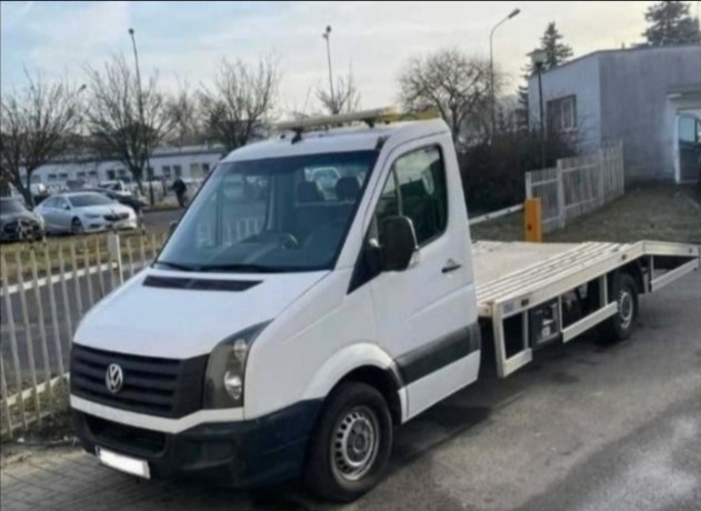 Biała laweta Volkswagen Crafter z platformą załadunkową na parkingu miejskim w pochmurny dzień. Widoczne detale pojazdu i otoczenie miejskie.