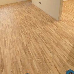 DREWPOL Cyklinowanie Podlog , Renowacja Schodow - Jasny parkiet ułożony w jodełkę w przestronnym, odświeżonym pomieszczeniu z białymi ścianami, widoczne niedoskonałości w postaci niezamaskowanego gniazdka elektrycznego i śladów po taśmie...