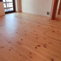 DREWPOL Cyklinowanie Podlog , Renowacja Schodow - Jasna, odnowiona podłoga z drewna sosnowego w przestronnym pokoju, widoczne sęki i naturalne usłojenie, z fragmentem schodów w tle.