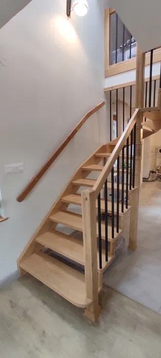 Jasne, drewniane schody z czarnymi balustradami i zakrzywionym poręczą na ścianie, oświetlone lampą sufitową, widok z dołu.