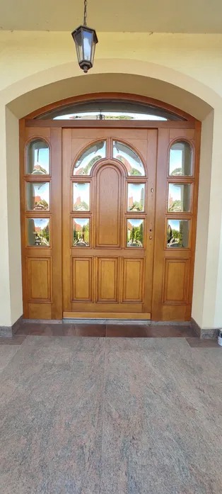 Drewniane drzwi wejściowe z szybkami, odnowione i polakierowane, z widocznym odbiciem ogrodu w szybach.