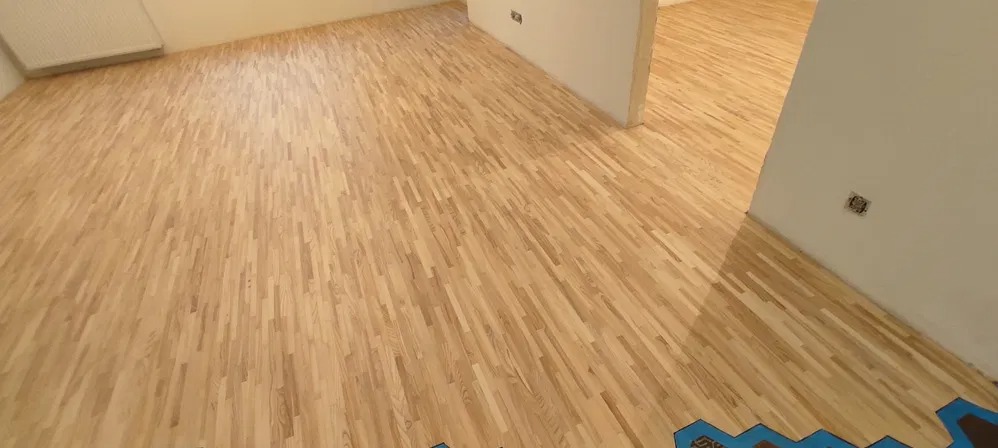 Jasny parkiet ułożony w jodełkę w przestronnym, odświeżonym pomieszczeniu z białymi ścianami, widoczne niedoskonałości w postaci niezamaskowanego gniazdka elektrycznego i śladów po taśmie...