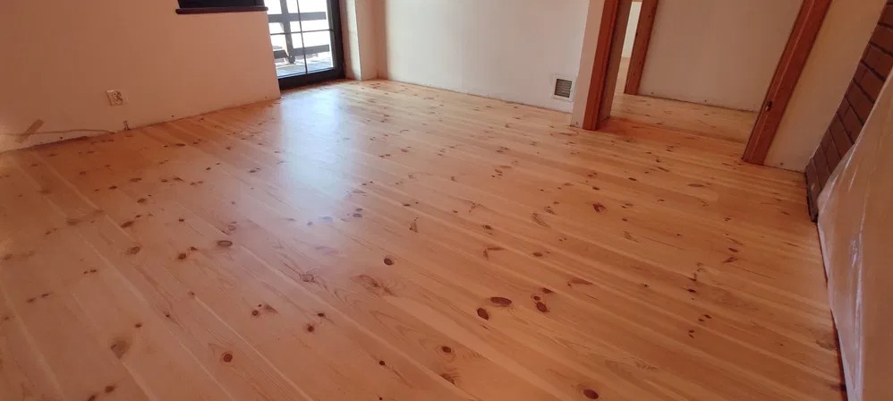 Jasna, odnowiona podłoga z drewna sosnowego w przestronnym pokoju, widoczne sęki i naturalne usłojenie, z fragmentem schodów w tle.